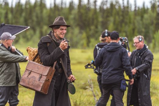 Skådespelaren Gustaf Skarsgård på en inspelningsplats iklädd gammeldags rock, väska, hatt och pipa.