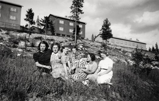 Sju personer sitter på marken och i bakgrunden är gruvbyn Laver på 1940-talet.