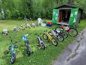 Bild på ett förråd och uppradade barncyklar på en gräsmatta.
