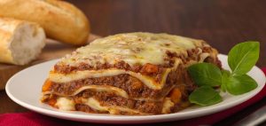 Bilden visar en vit tallrik med lasagne och en grön kvist med basilika.