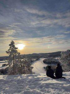Bilden visar Piteälven sett från ett berg. På berget sitter två personer som tittar ut över älven. Det är vinter med en sol bakgrunden.