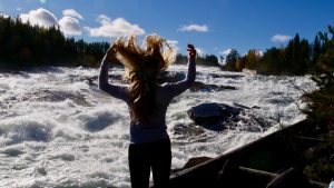 Bilden visar en kvinnlig person bredvid Storforsen. Personen har armarna utsträckta uppåt och håret spretar uppåt.