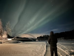 Bilden visar en person som vandrar på en frusen älv, på väg mot en bro under ett norrsken.